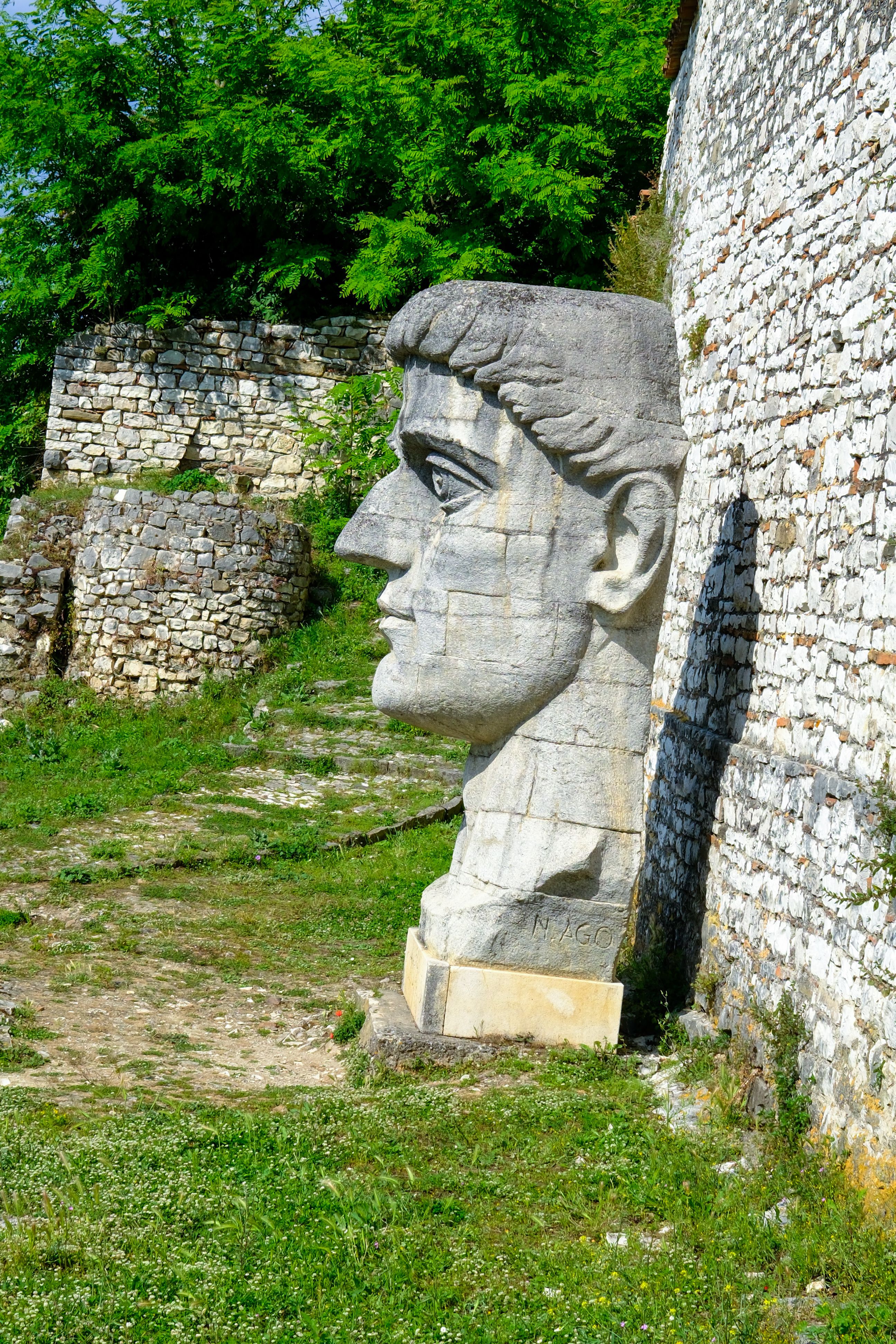 Stone head, Roman style sculpture near old stone wall surrounded by greenery.