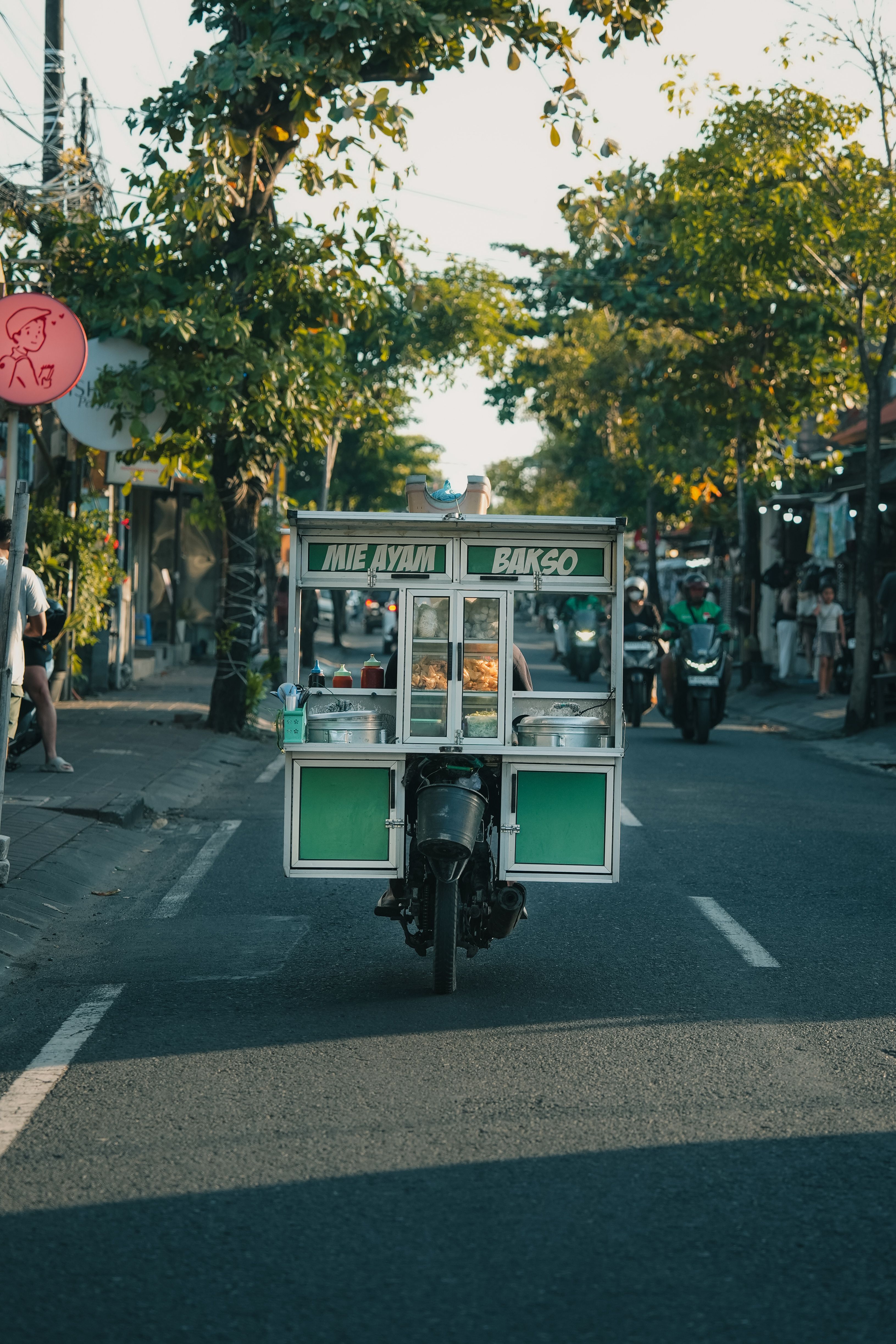 A mobile food cart serves Mie Ayam Bakso on a busy city street lined with trees.