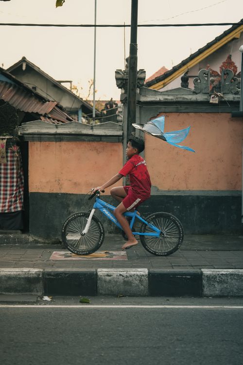 Free Young boy riding a bicycle with a kite in an urban Asian neighborhood, showcasing daily life. Stock Photo