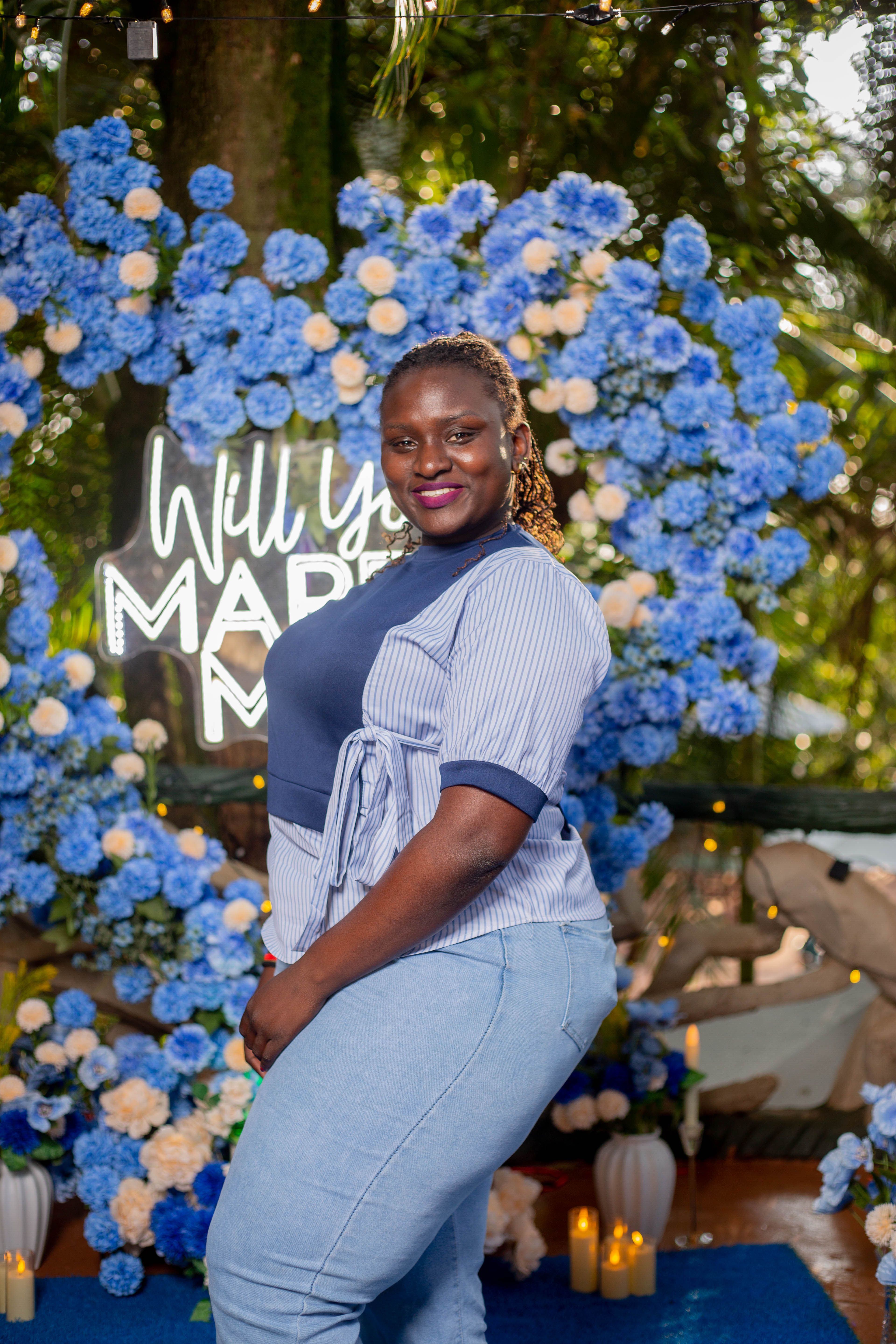 Smiling woman in front of a 'Will You Marry Me?' floral heart backdrop.