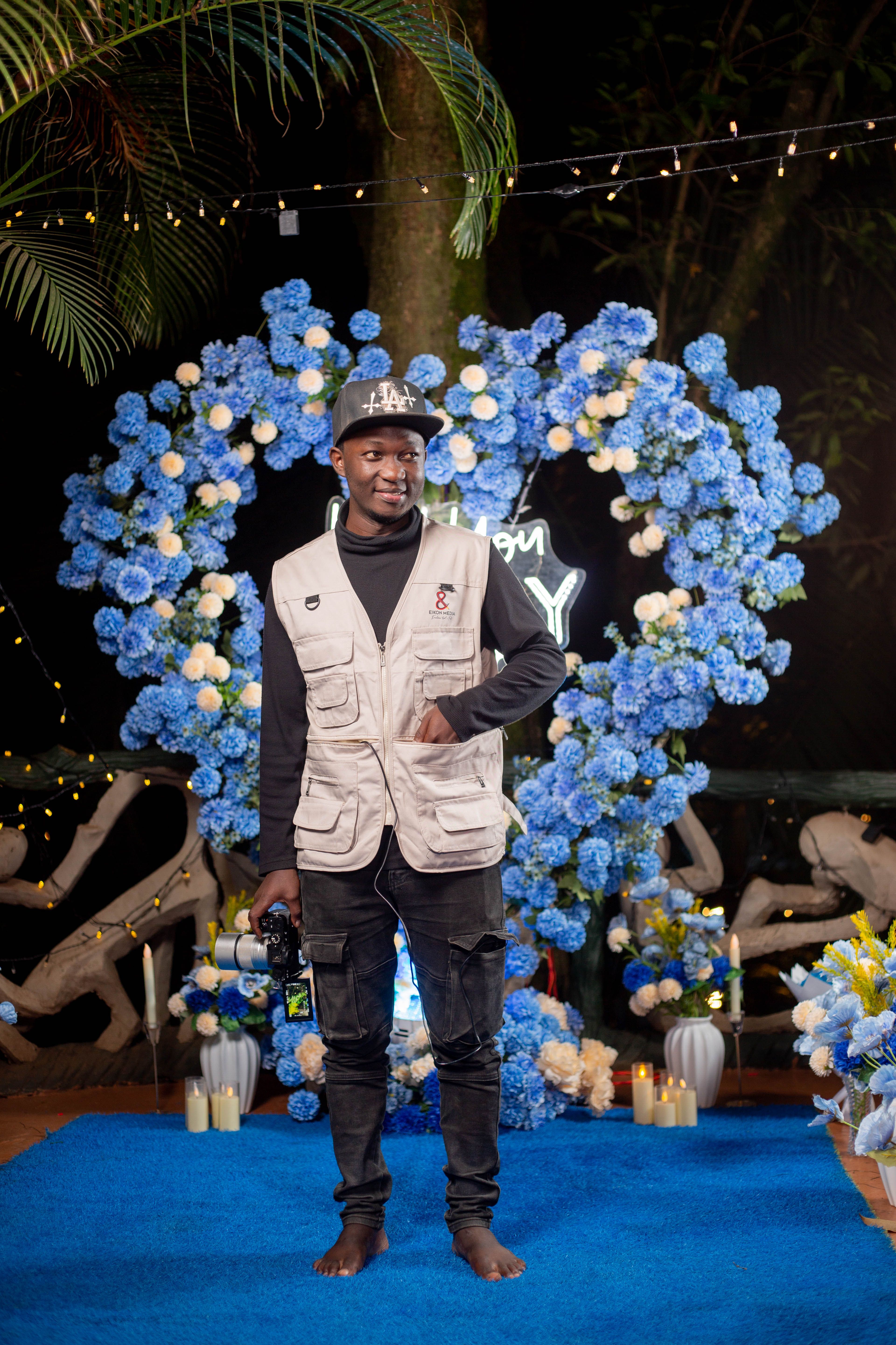 Person posing with a camera at an outdoor night event with blue floral decorations.