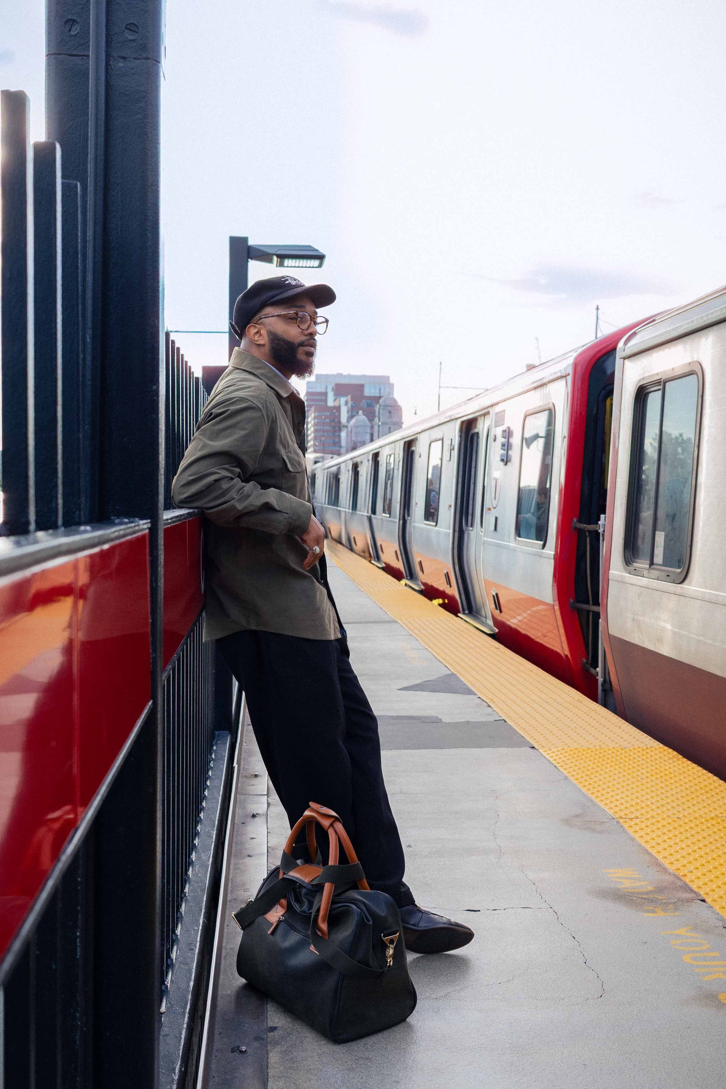 Man in casual attire leans against railing at Boston subway, train arriving.