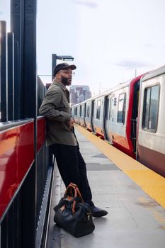 Man in casual attire leans against railing at Boston subway, train arriving.