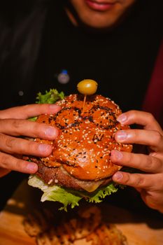 Close-up of hands holding a gourmet burger with a sesame seed bun and olive on top.