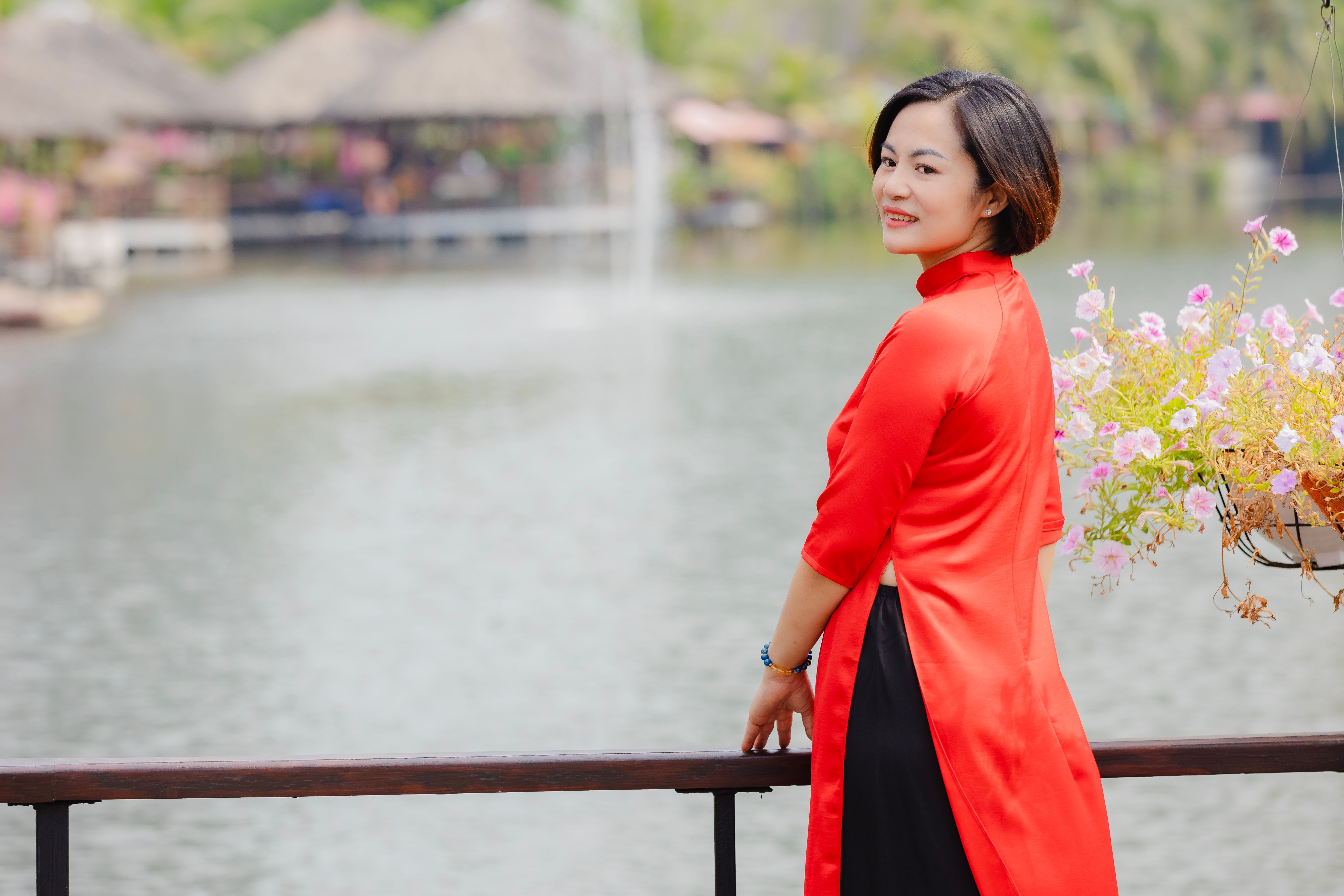 A woman in a red traditional dress stands by a lake, radiating elegance and serenity.