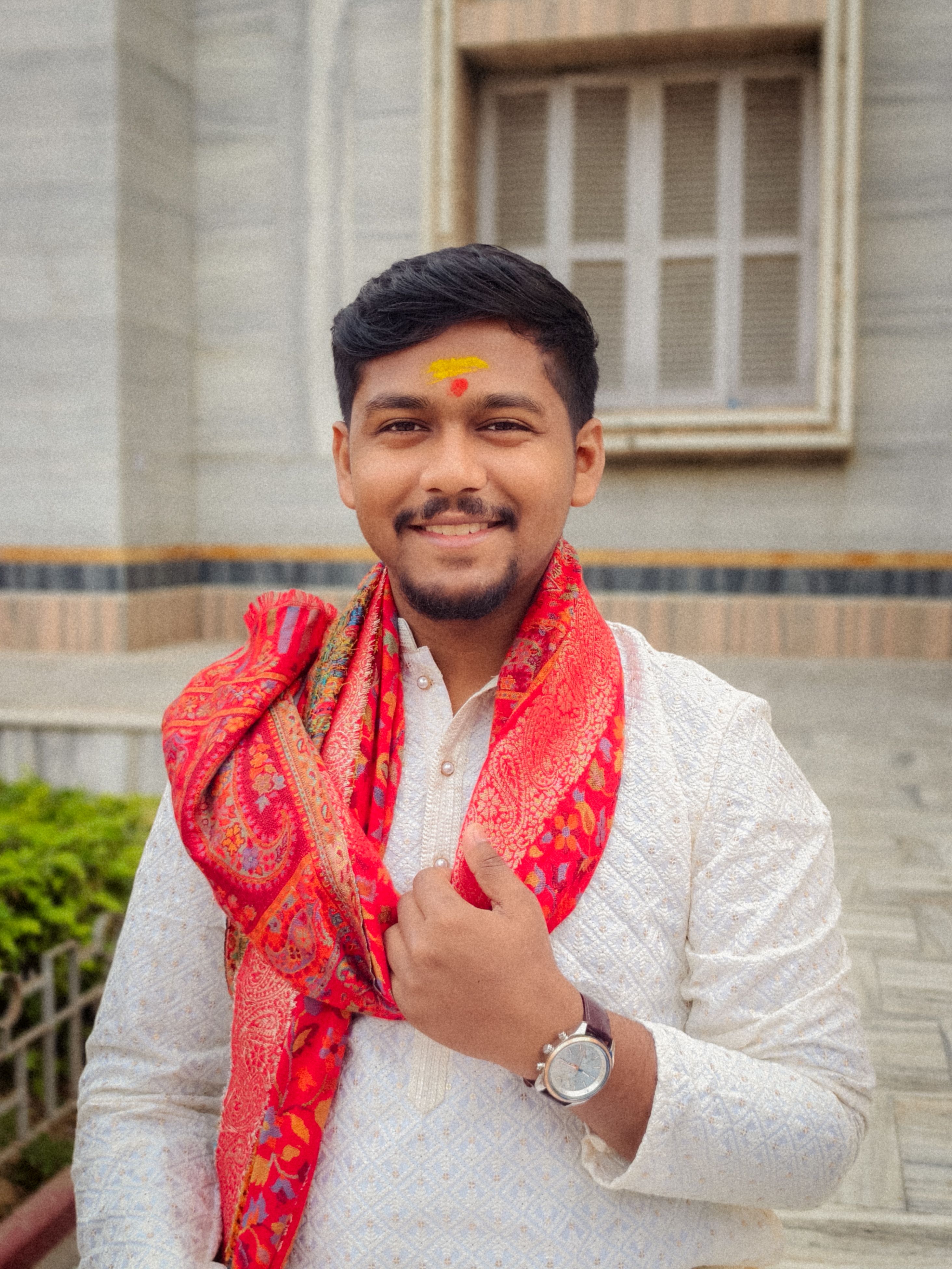 A young man with a red scarf and traditional clothing smiling outdoors.