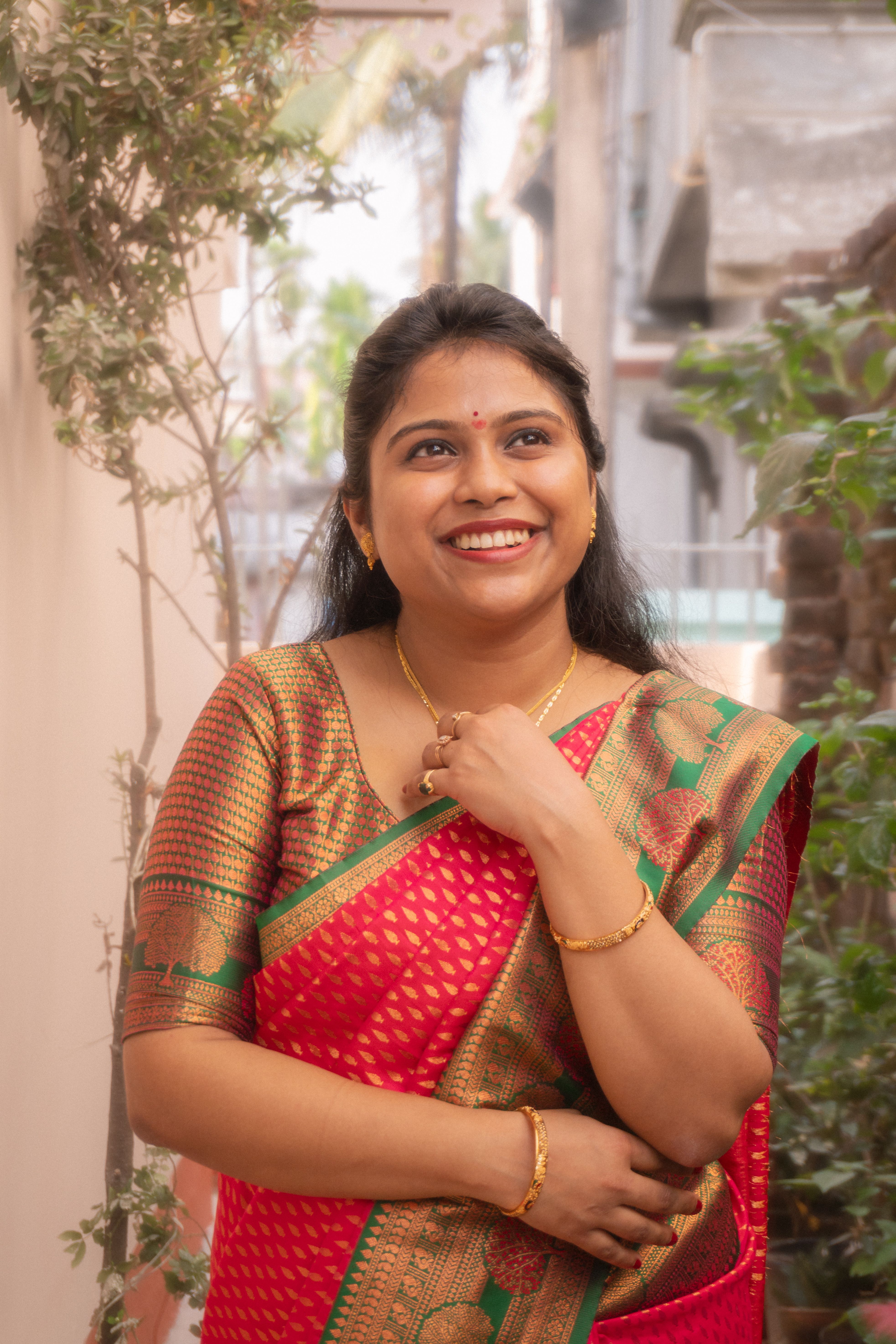 A joyful woman in a colorful saree smiles in an outdoor setting with foliage.