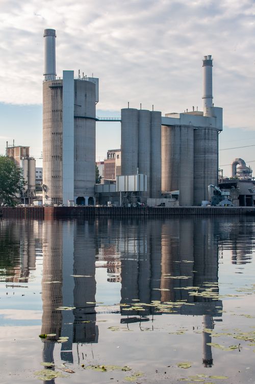 Free A large industrial plant's reflection is visible in a calm river, under a partly cloudy sky. Stock Photo