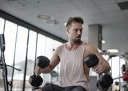 From below of muscular male pumping biceps with dumbbells while sitting on bench in sports center