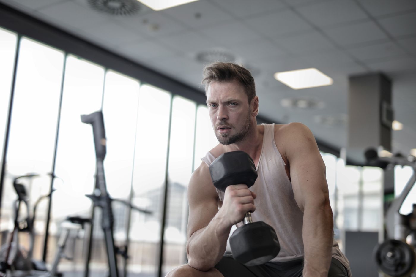 A lifter rests a dumbbell on his knee between sets beside a wall of windows