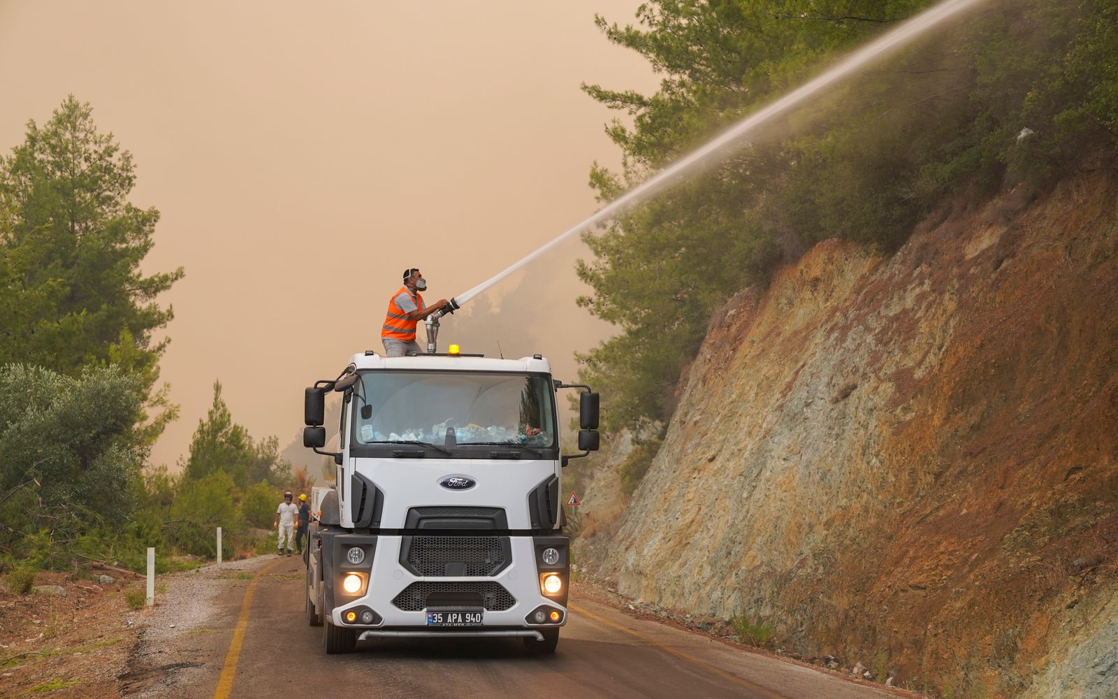 Firefighters in protective gear use a water cannon on a truck to fight a forest wildfire