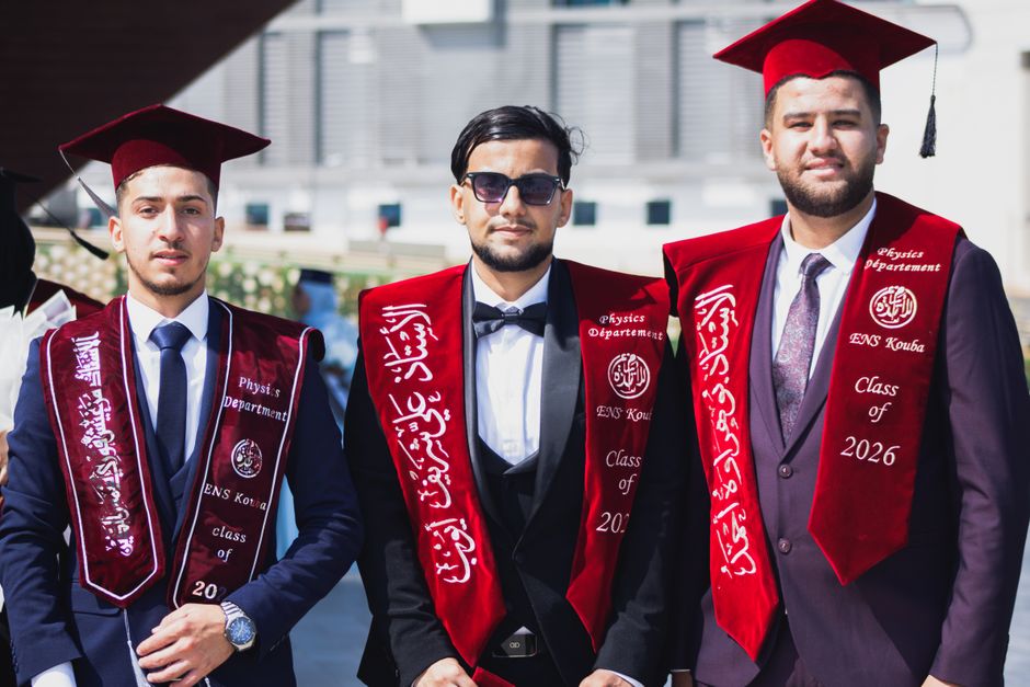 Three male graduates from ENS Kouba in Algiers, wearing caps and gowns, excitedly pose for a photo.