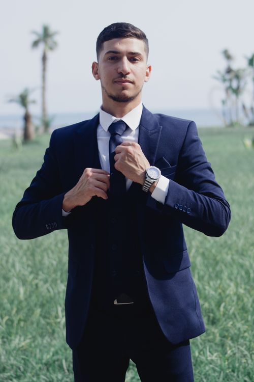 Free Young man adjusting tie in a formal suit, standing outdoors with palm trees in the background. Stock Photo