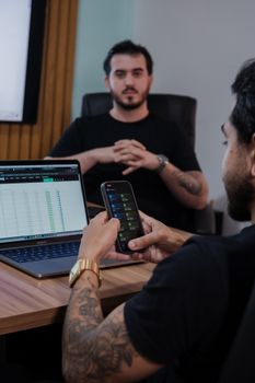 Two men in a modern office reviewing stock market data on laptop and smartphone.