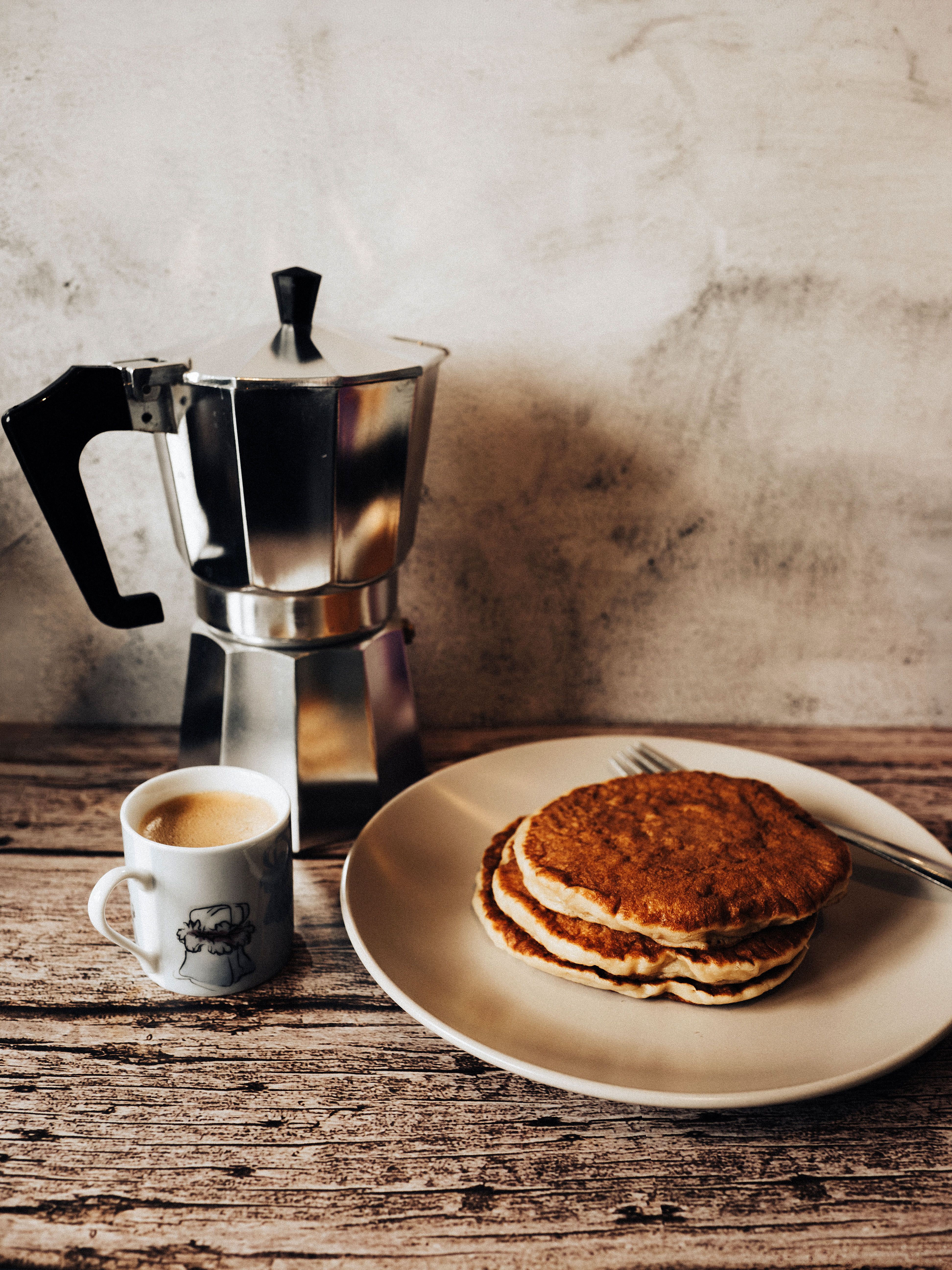 Rustic breakfast scene with a moka pot, espresso, and pancakes on a wooden table.