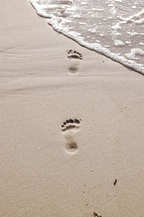 Free Close-up of human footprints on a sandy beach with ocean waves approaching. Stock Photo