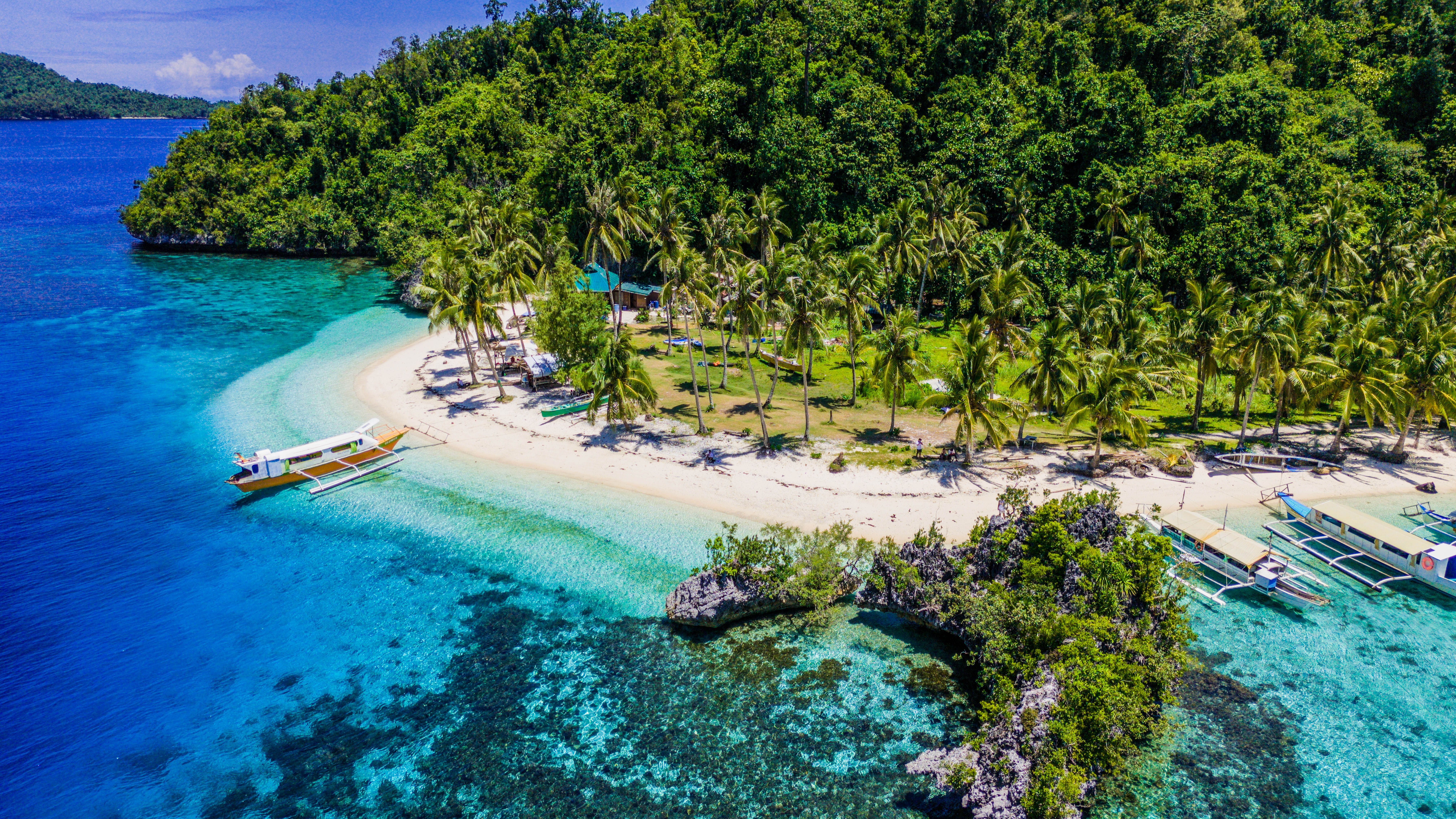 Aerial view of a tropical beach in the Philippines with outrigger boats anchored in clear turquoise waters near palm trees.
