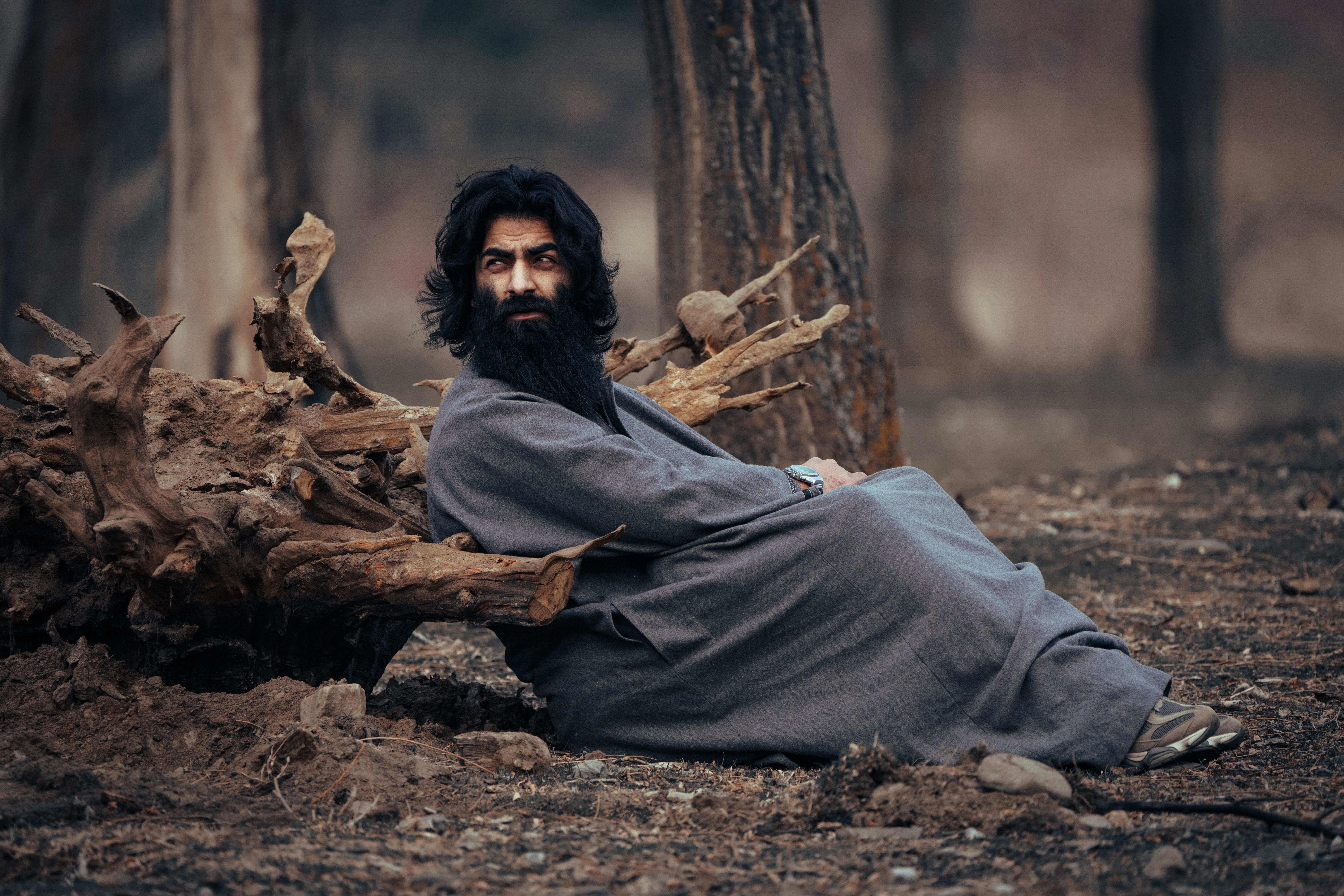Fotomural Un hombre barbudo con vestimenta tradicional descansa apoyado en la raíz de un árbol en un entorno forestal sereno.