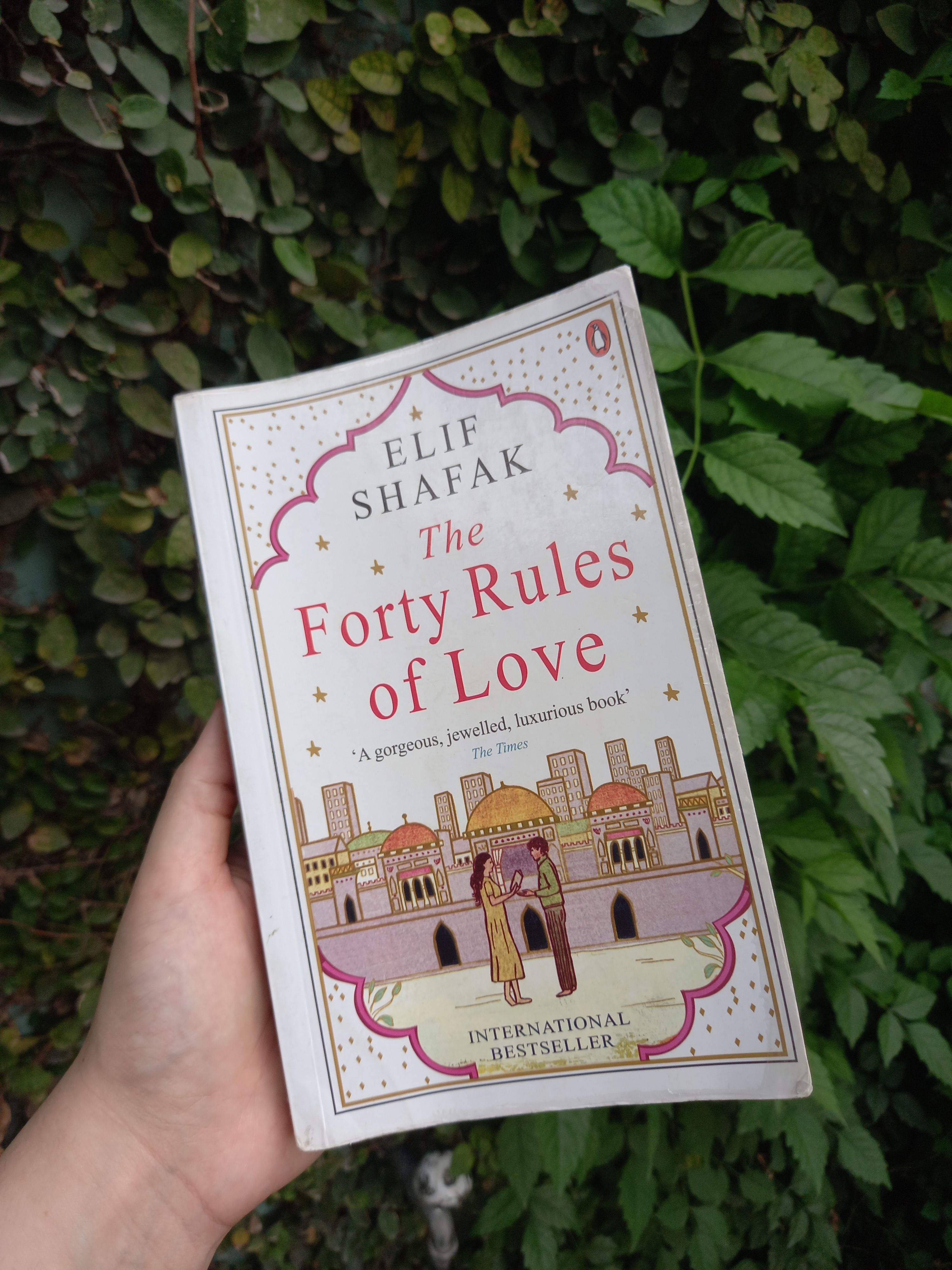 Person outdoor holding a book 'The Forty Rules of Love' with greenery in background.