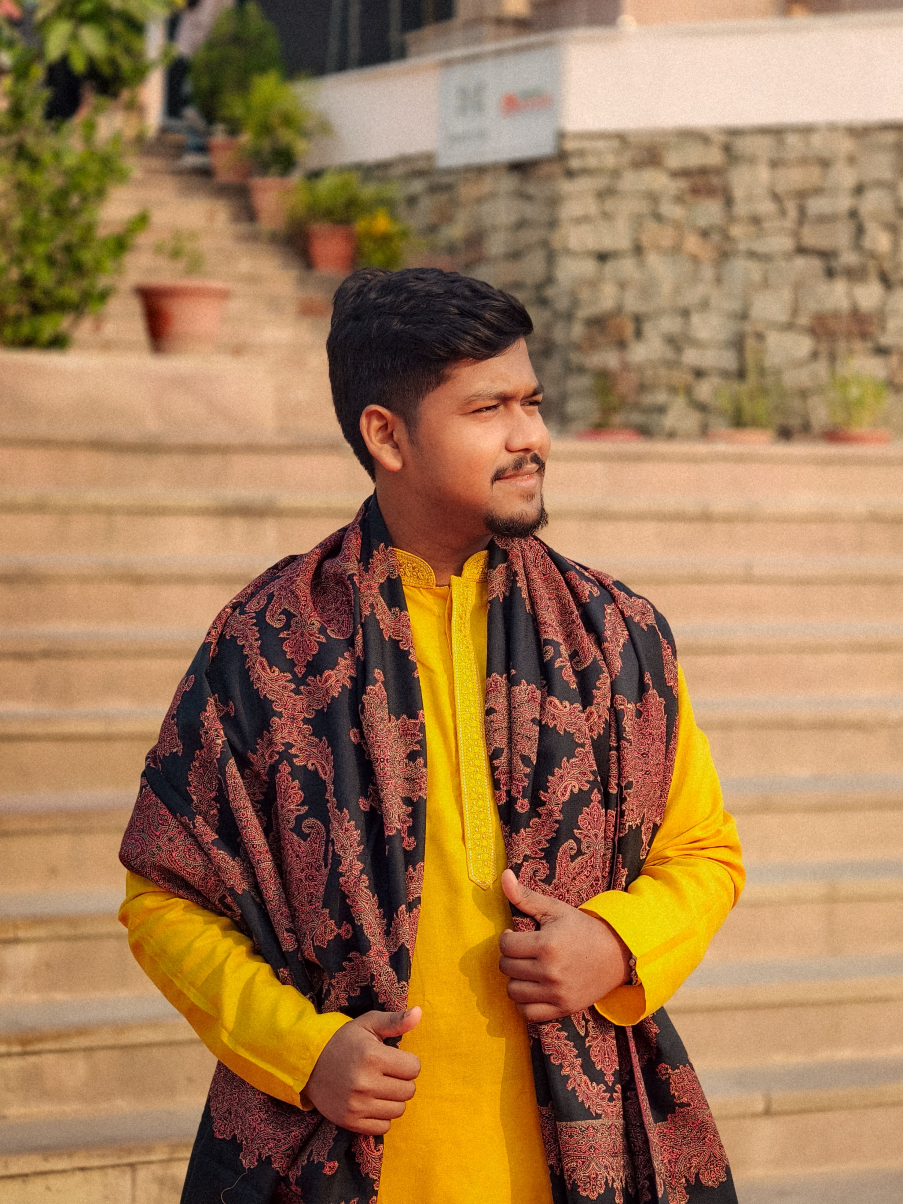 An adult male in a yellow kurta and patterned shawl stands confidently on outdoor stone steps.