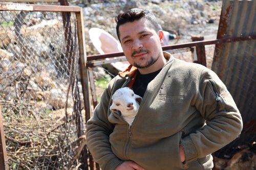 Free Smiling man holding a baby goat in a rustic outdoor farm area. Stock Photo