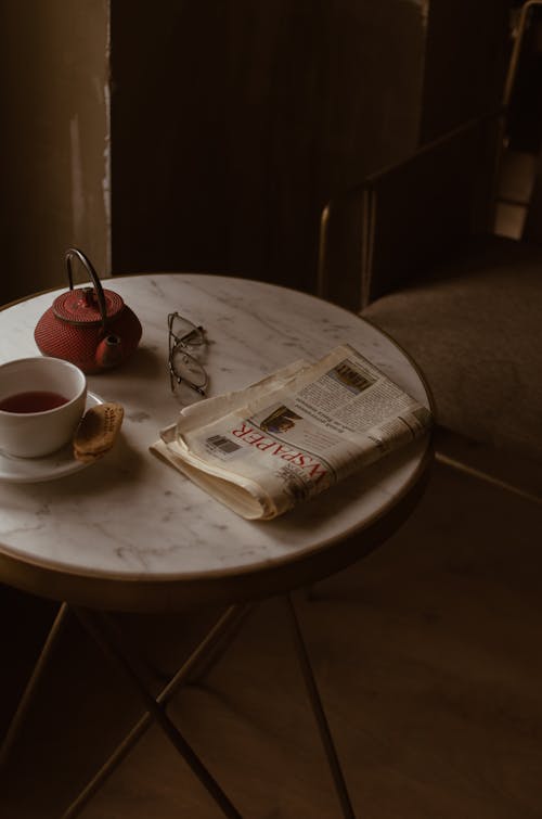 From Above Of Round Marble Textured Table With Tea Set Placed Near Newspaper And Eyeglasses In Retro Style Cafe