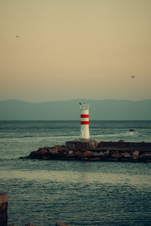 Free A serene view of a red and white lighthouse at twilight by the sea with mountains in the background. Stock Photo