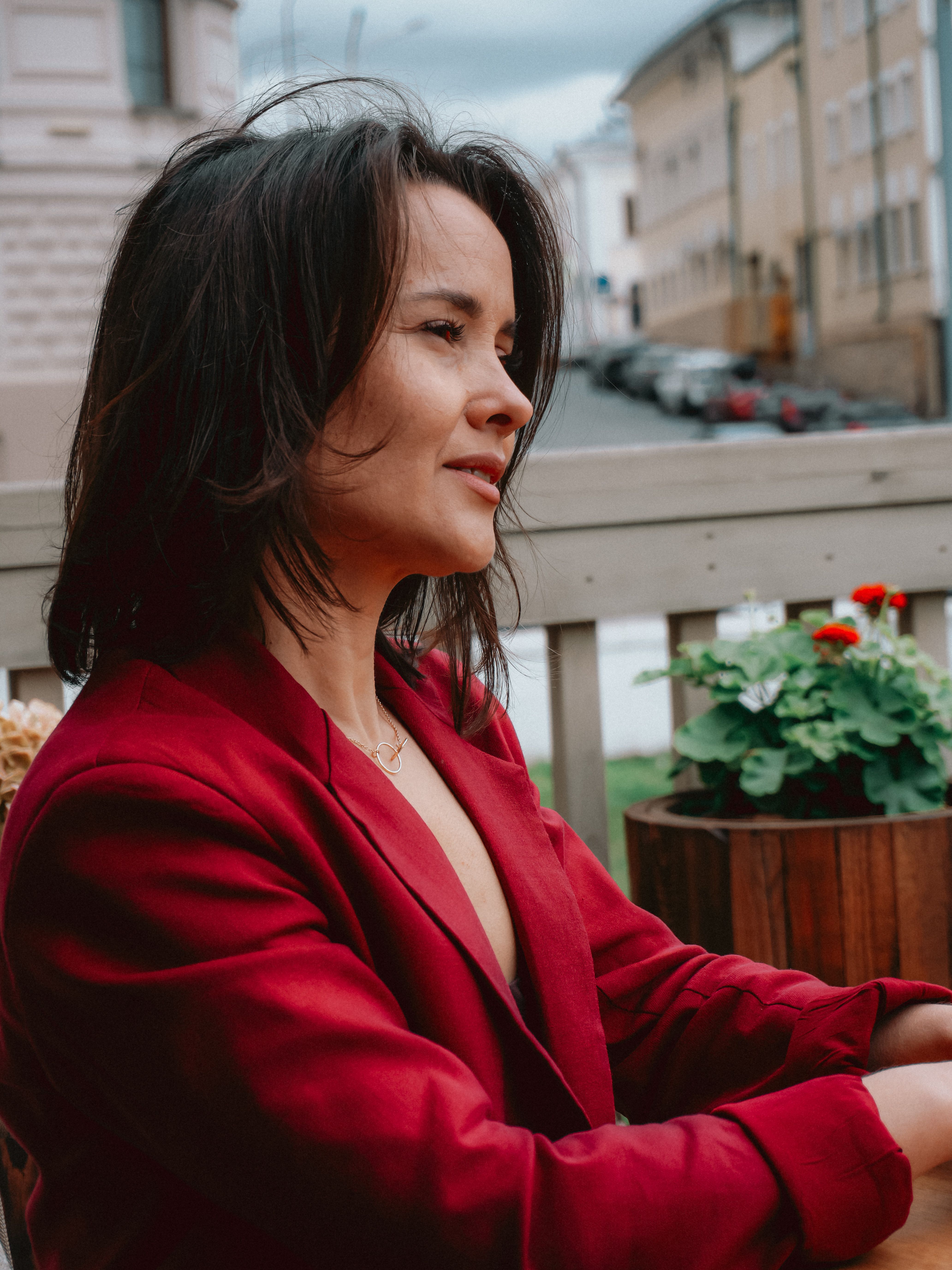 A woman in a red blazer enjoys a relaxed moment outdoors on a terrace with a cityscape view.