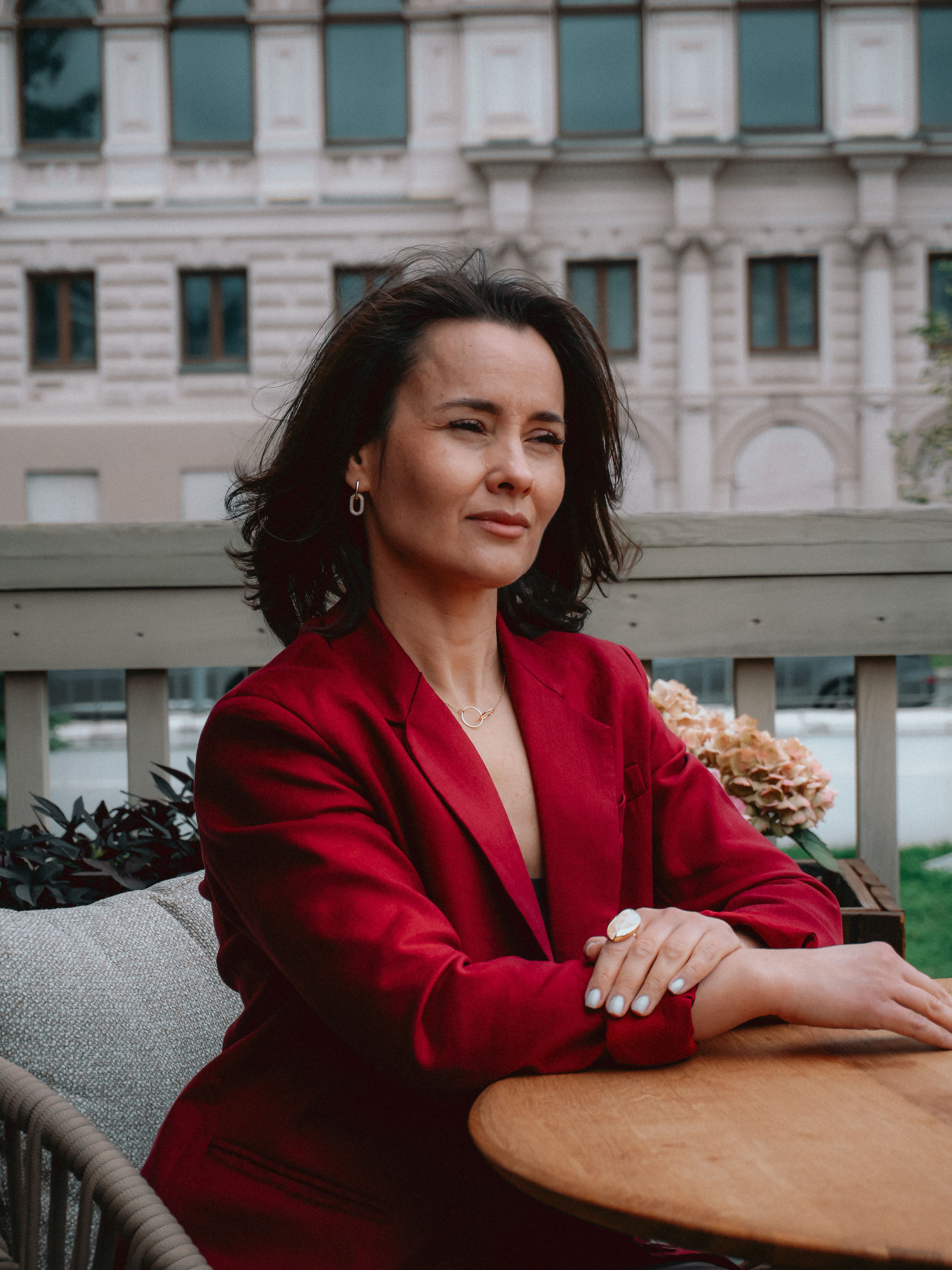 Elegant woman in red suit sits at outdoor terrace table, gazing thoughtfully.