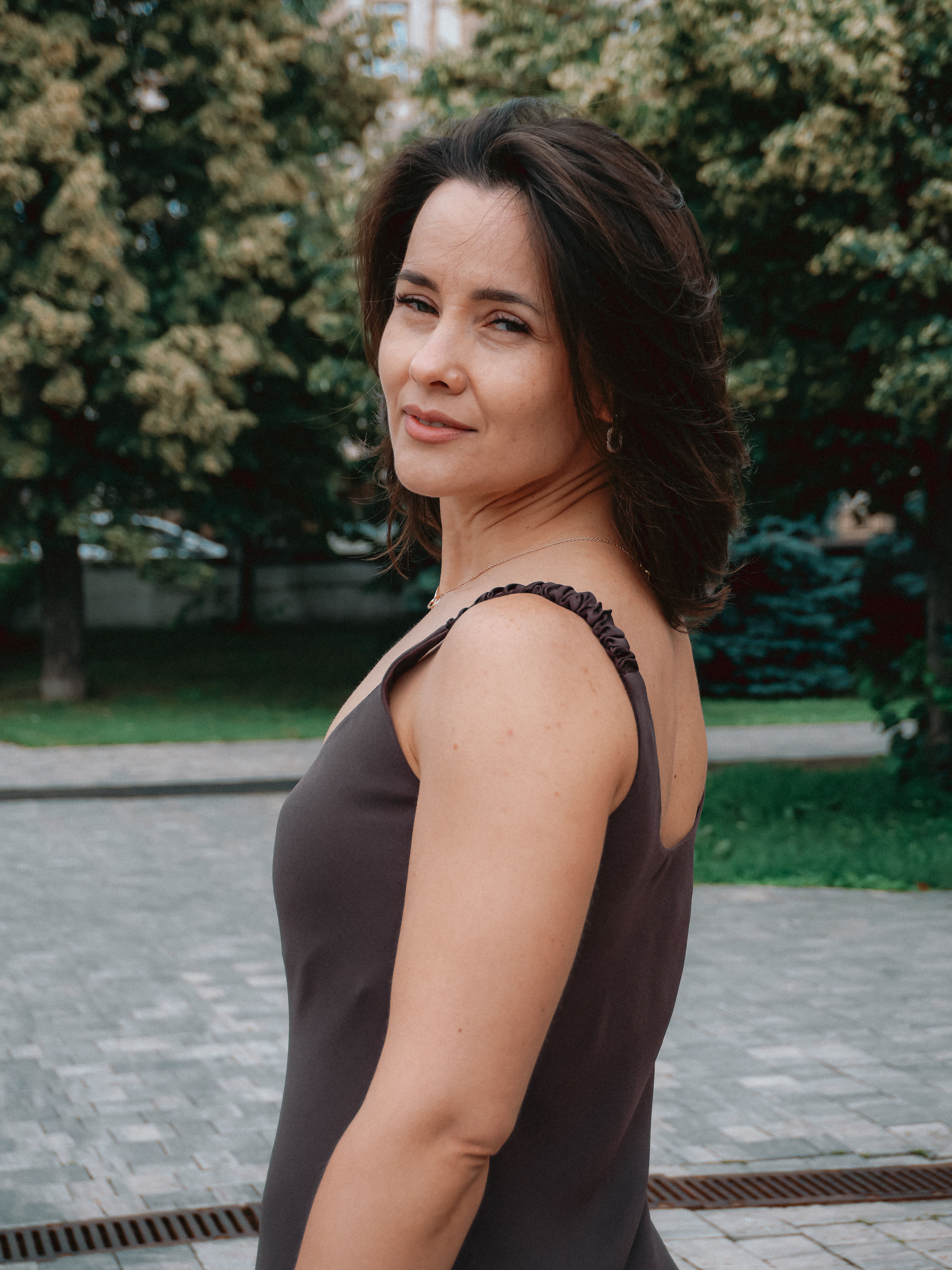 A confident woman in casual attire poses in a serene outdoor setting, embodying poise and grace.