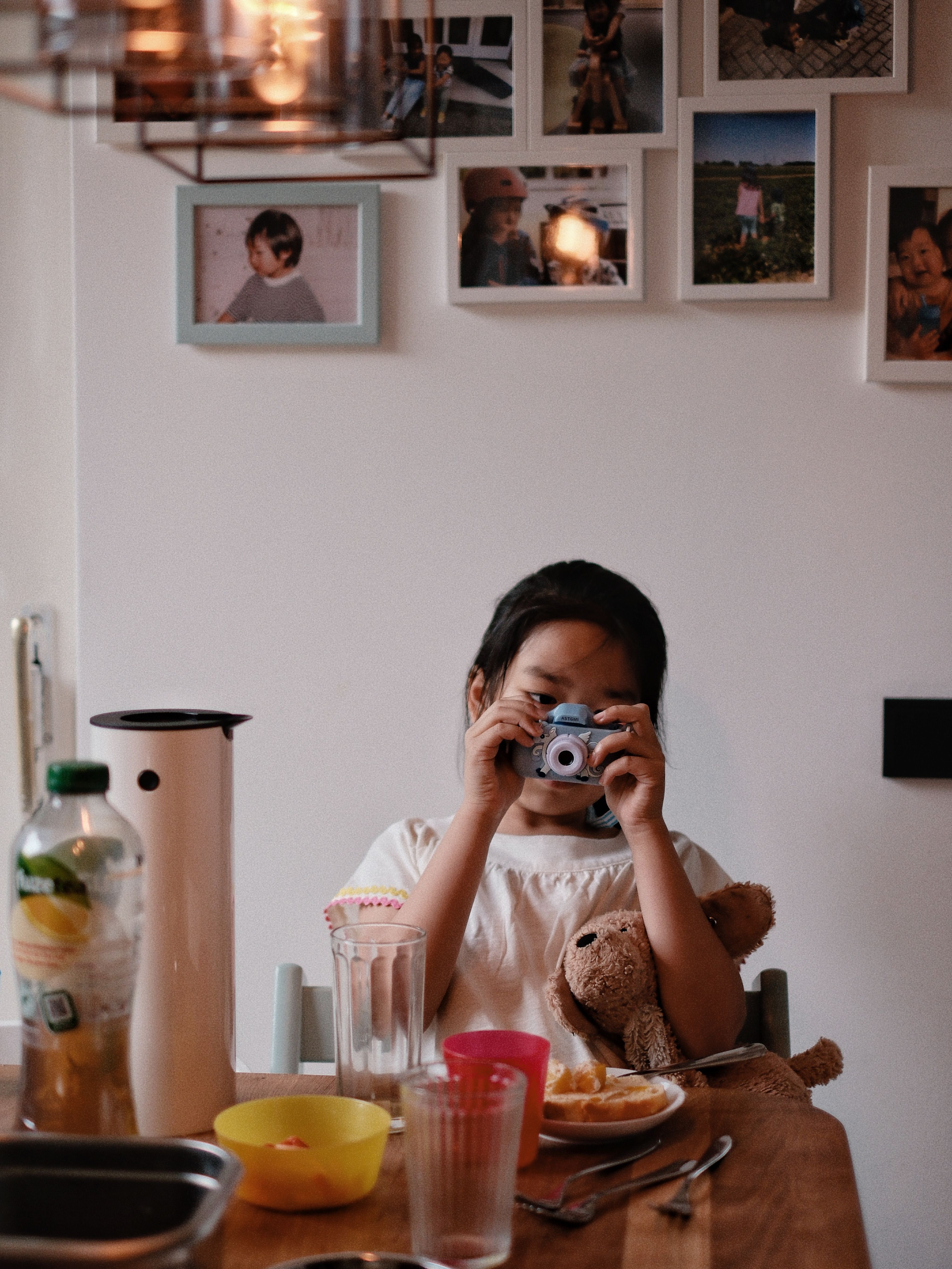 A young girl enjoys breakfast with a toy camera and teddy bear, set in a cozy kitchen.