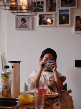 A young girl enjoys breakfast with a toy camera and teddy bear, set in a cozy kitchen.