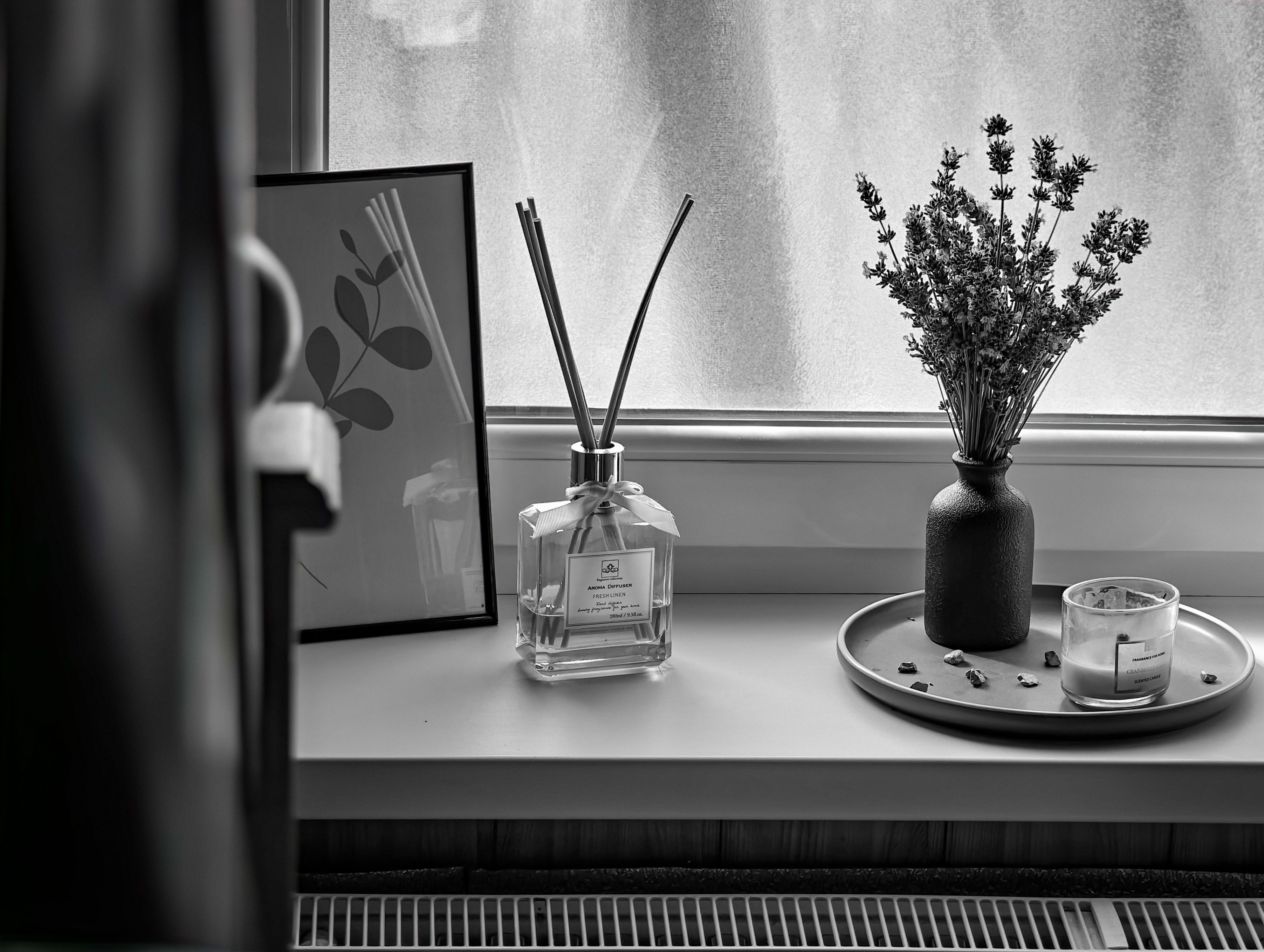 Black and white image of a windowsill with reed diffuser, lavender, and candle.