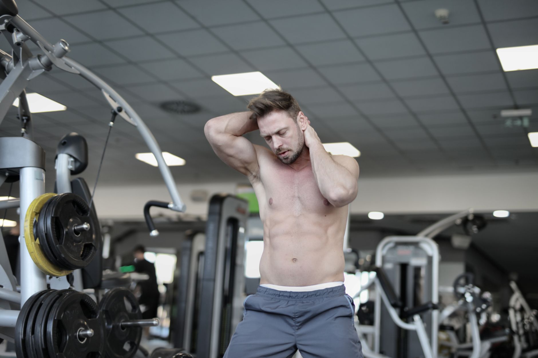 Muscular shirtless sportsman warming up neck muscles with hands standing with closed eyes in modern sport club near weightlifting equipment