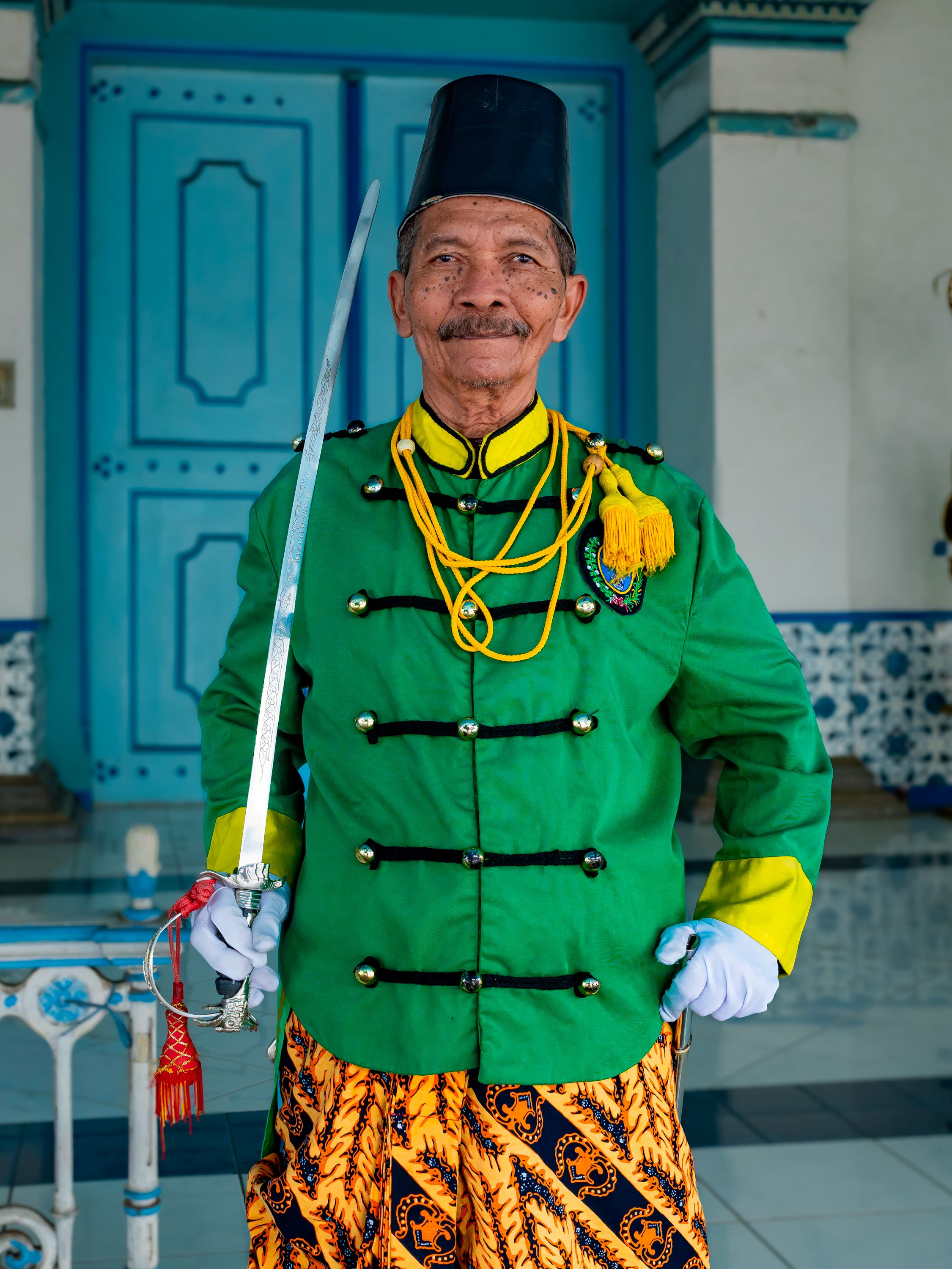 Elderly Javanese man in vibrant cultural attire holding a sword in a historic Indonesian setting.