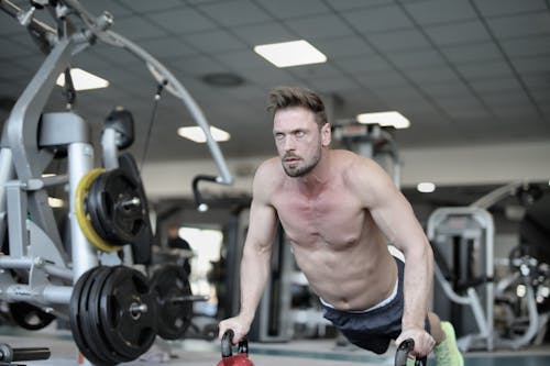 Free Concentrate muscular sportsman with naked torso performing push ups on kettlebells and looking away while training in modern fitness club Stock Photo