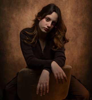 A contemplative portrait of a young woman seated on a chair against a warm, dark background.