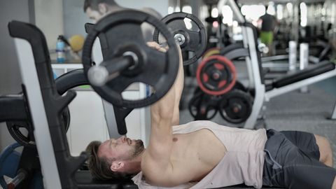 Side view of strong young sportsman in sportswear lying on bench and pressing heavy barbell doing chest exercise training in modern gym