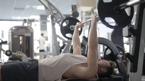 Side view of strong young sportsman in casual sportswear pressing heavy barbell on bench while intense training in modern fitness center