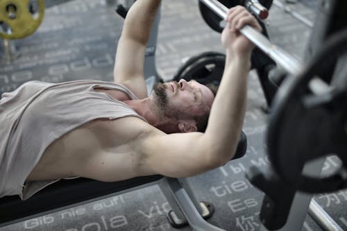 From above view of strong bearded sportsman in sportive tank top concentrating during bench press with barbell while training in modern fitness club