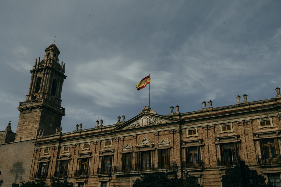 Arquitectura histórica en Valencia con la bandera española ondeando. Un cielo dramático realza la escena.