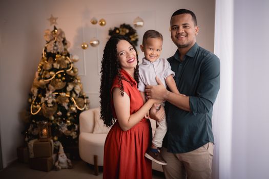A smiling family of three poses joyfully by a decorated Christmas tree indoors.