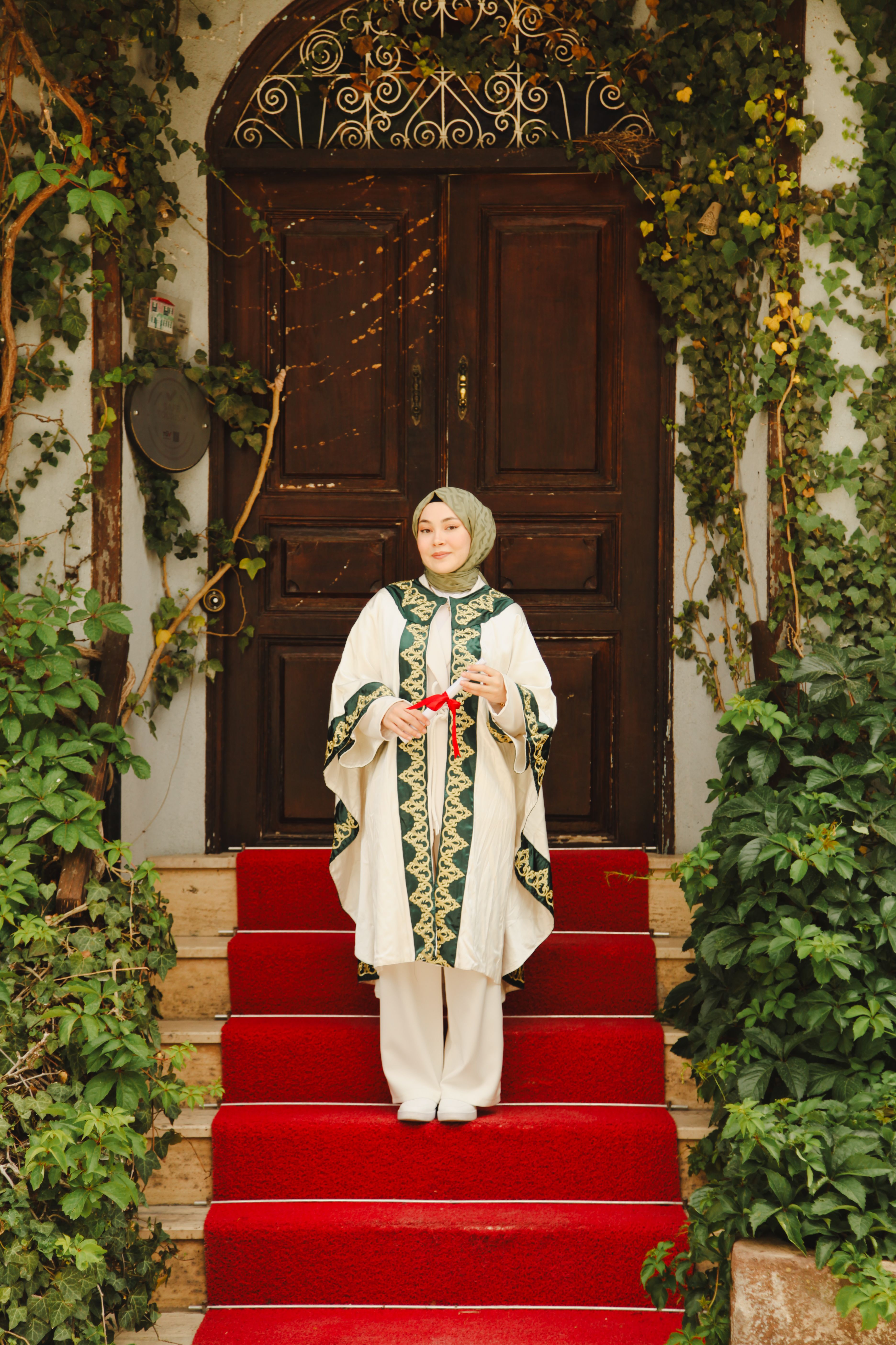 A woman in traditional attire holds a gift on steps with a red carpet and lush greenery.