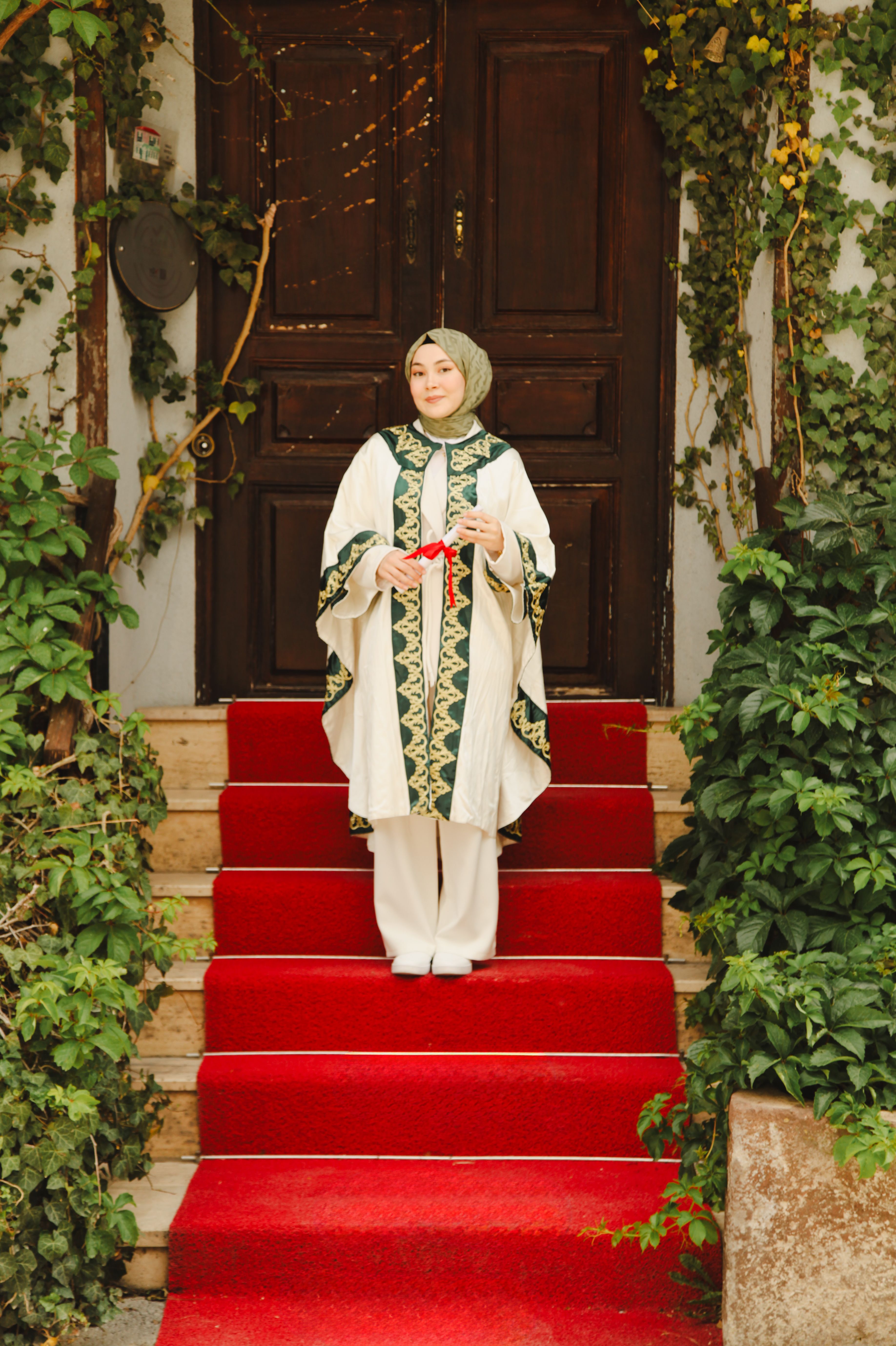 Woman in embroidered attire with headscarf standing on a red carpeted staircase outside a wooden door.