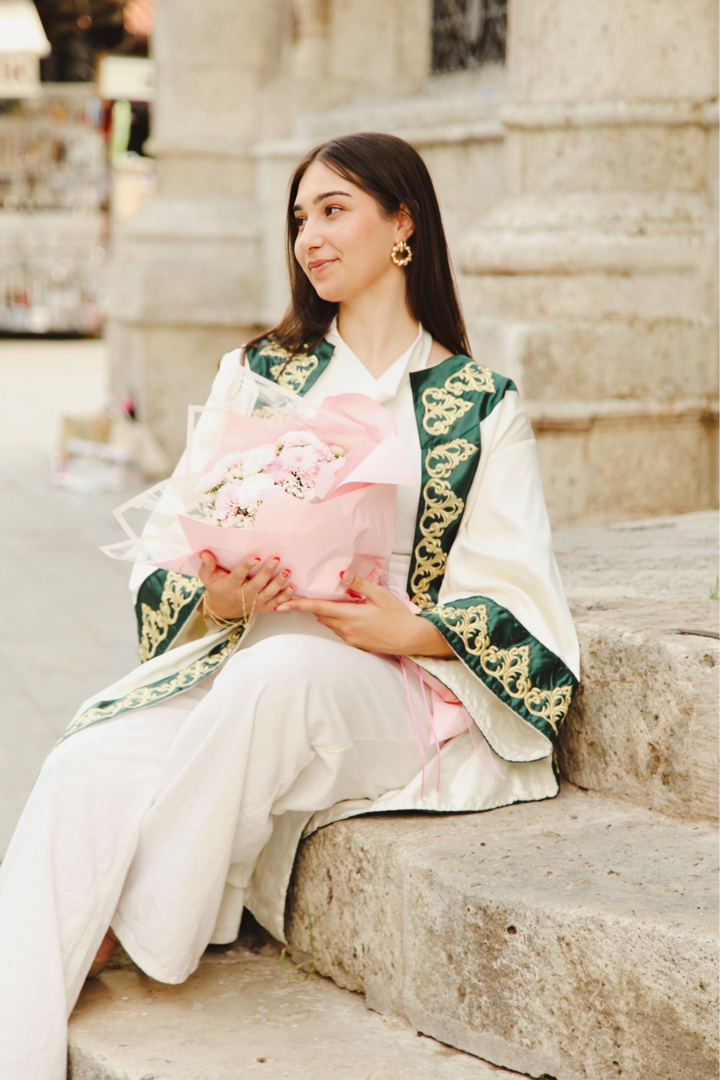 Graduating student in ceremonial outfit holding bouquet, sitting outdoors.