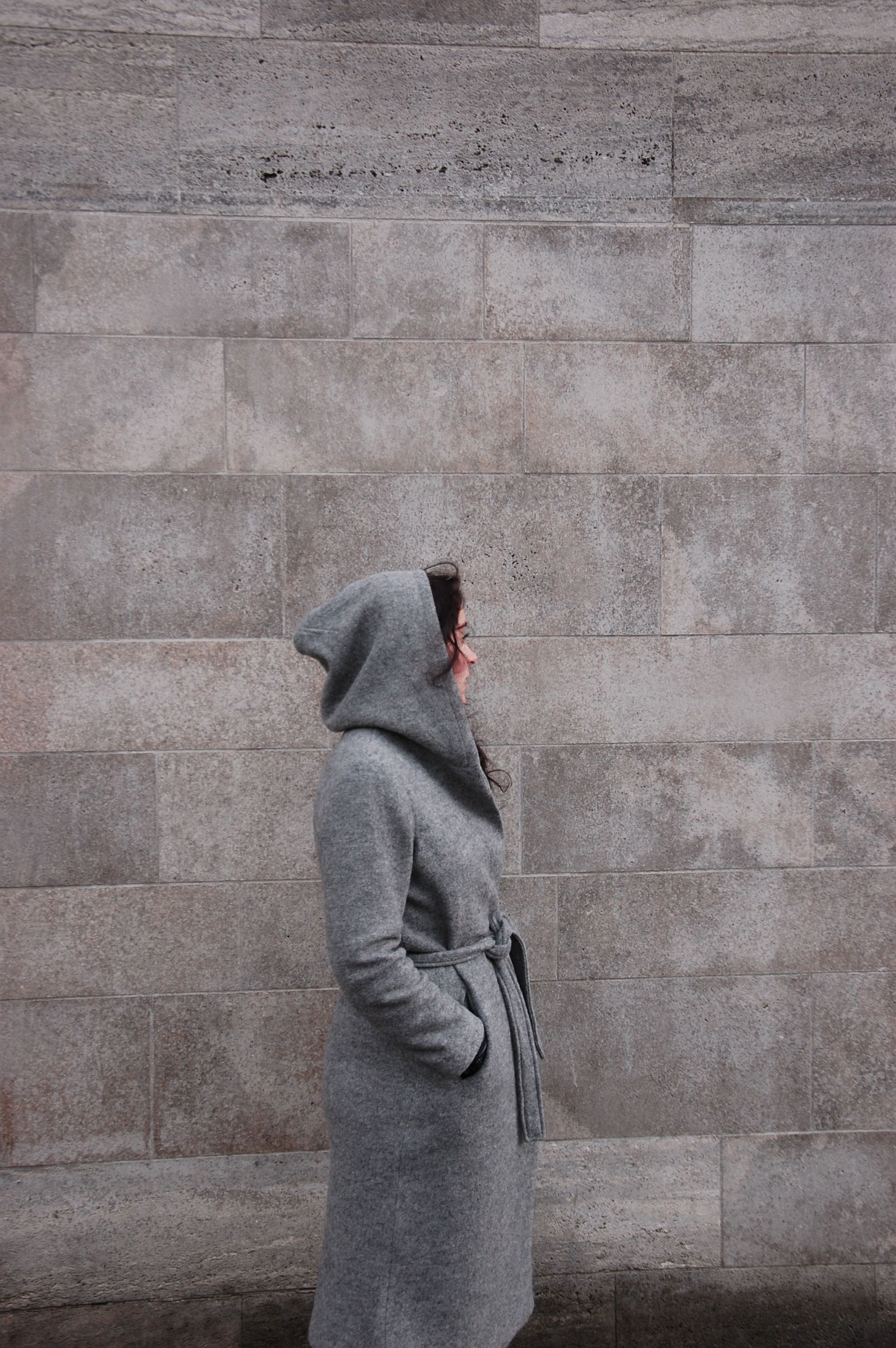 Side profile of a woman in a hooded grey coat standing against a textured stone wall.