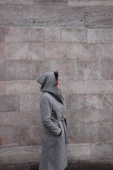 Side profile of a woman in a hooded grey coat standing against a textured stone wall.
