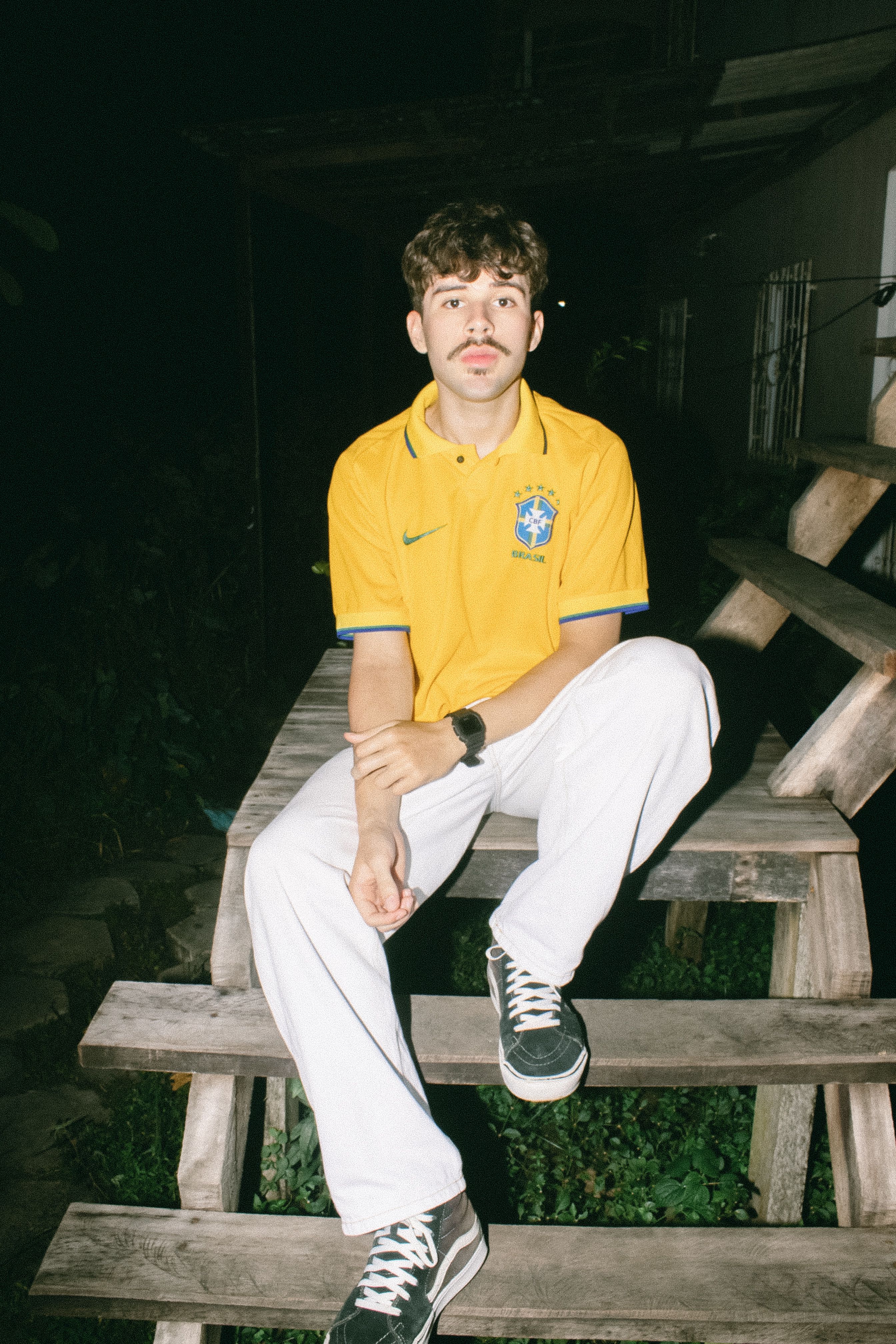 Young man wearing a Brazil soccer jersey, sitting on stairs at night. Casual, sporty ambiance.