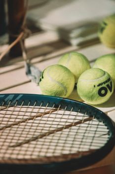 Focused shot of a tennis racket and several tennis balls on an outdoor court bench.