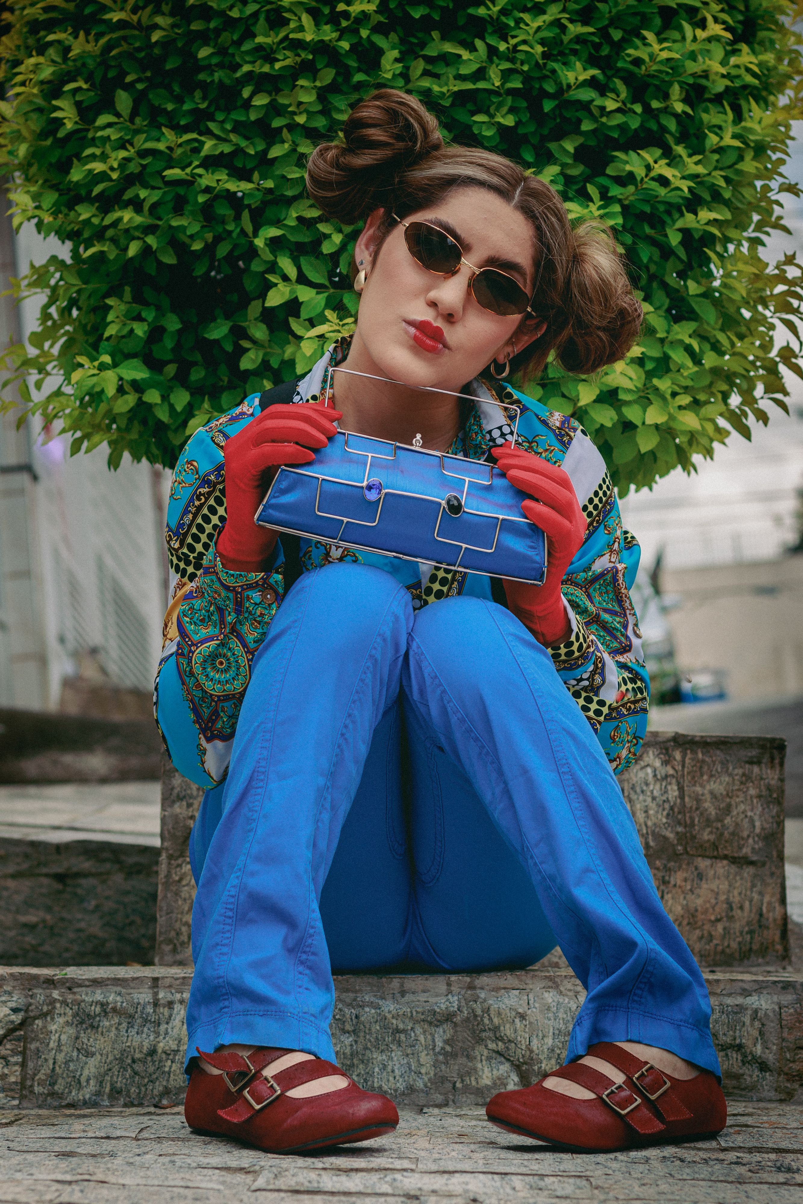 Um retrato marcante de uma mulher com roupas vintage coloridas, posando ao ar livre com óculos de sol e uma bolsa vibrante.